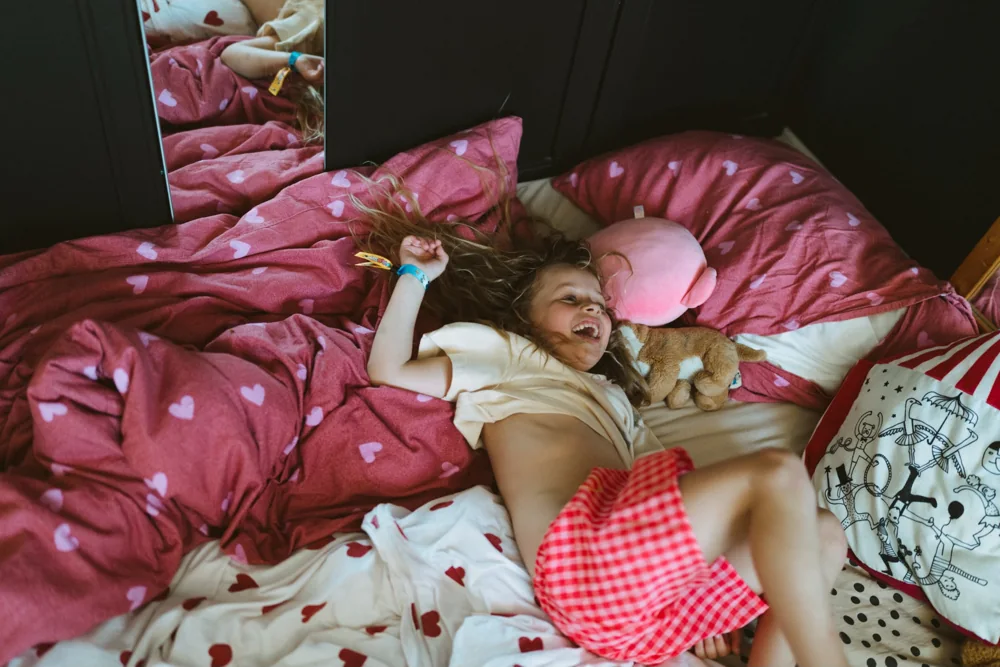 Een meisje ligt te lachen in een onopgemaakt bed, haar haar alle kanten op